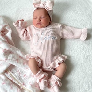 Puede incluir: Un beb&eacute; reci&eacute;n nacido con un conjunto de punto rosa claro, que incluye un su&eacute;ter con el nombre "Clara" bordado en la parte delantera, bragas con volantes a juego, calcetines y una diadema con lazo. El beb&eacute; est&aacute; acostado sobre una superficie blanca.