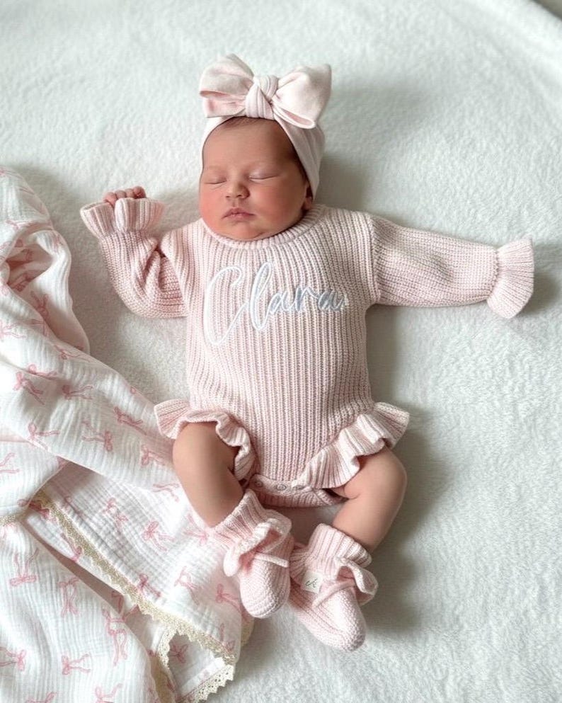 Puede incluir: Un beb&eacute; reci&eacute;n nacido con un conjunto de punto rosa claro, que incluye un su&eacute;ter con el nombre "Clara" bordado en la parte delantera, bragas con volantes a juego, calcetines y una diadema con lazo. El beb&eacute; est&aacute; acostado sobre una superficie blanca.