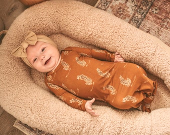 Könnte beinhalten: Ein Baby trägt ein braunes und beigefarbenes, geknotetes Kleid mit Leopardenmuster. Das Baby liegt auf dem Rücken in einem cremefarbenen, flauschigen Nest. Das Baby trägt ein braunes Schleifenband.