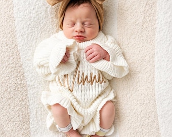 May include: A cream-colored knit romper with long sleeves and ruffled edges, featuring the embroidered name Winnie in gold thread across the chest. The garment is paired with a matching tan fabric bow headband.