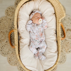 May include: A baby wearing a floral print sleeper and a headband is sleeping in a woven basket with a white padded liner.
