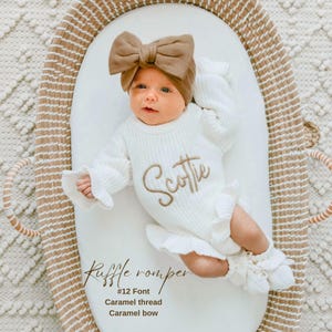 May include: A white long-sleeve knit romper with ruffled edges and the name Scottie embroidered in caramel-colored thread across the chest. The outfit includes a matching caramel-colored fabric bow headband and knit booties.