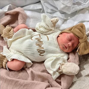 Puede incluir: Un reci&eacute;n nacido durmiendo con un conjunto de punto color crema con el nombre "Emma" bordado en dorado. El conjunto incluye un gorro y manoplas a juego. El beb&eacute; est&aacute; sobre una manta rosa, con una diadema de lazo color canela.
