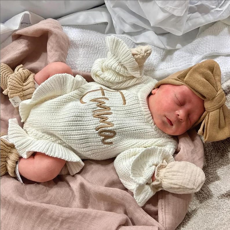 Puede incluir: Un reci&eacute;n nacido durmiendo con un conjunto de punto color crema con el nombre "Emma" bordado en dorado. El conjunto incluye un gorro y manoplas a juego. El beb&eacute; est&aacute; sobre una manta rosa, con una diadema de lazo color canela.