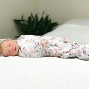 May include: A baby swaddled in a white and pink floral swaddle with a matching headband. The baby is sleeping peacefully on a white bed.
