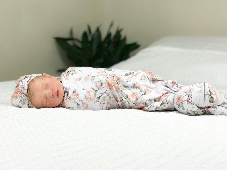 May include: A baby swaddled in a white and pink floral swaddle with a matching headband. The baby is sleeping peacefully on a white bed.
