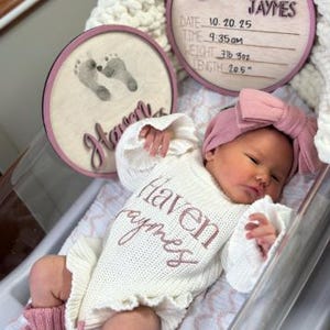 Puede incluir: Un beb&eacute; reci&eacute;n nacido con un body de punto blanco con el nombre "Haven Jaymes" bordado en rosa. El beb&eacute; tambi&eacute;n lleva una diadema rosa y calentadores a juego. Dos placas circulares de madera con el nombre "Haven Jaymes" y huellas de beb&eacute; en el fondo.