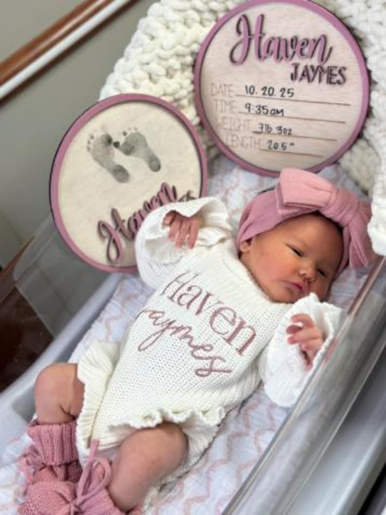 Puede incluir: Un beb&eacute; reci&eacute;n nacido con un body de punto blanco con el nombre "Haven Jaymes" bordado en rosa. El beb&eacute; tambi&eacute;n lleva una diadema rosa y calentadores a juego. Dos placas circulares de madera con el nombre "Haven Jaymes" y huellas de beb&eacute; en el fondo.