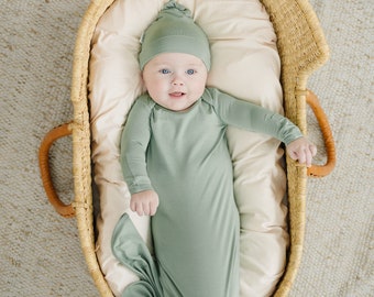 May include: A baby wearing a sage green knotted gown and matching hat is lying in a wicker basket. The basket is on a white rug.