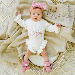 May include: A newborn baby wearing a white knit ruffle romper with the name "Haizley" embroidered in pink. The baby also wears a pink bow headband and matching pink knit booties. The words "NEW! Ruffle romper" are written in brown script.