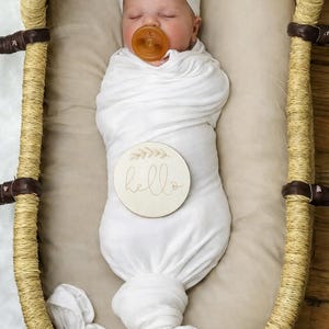 May include: A swaddled infant wearing a white headband and a brown pacifier, lying in a woven bassinet. A round wooden disc rests on the swaddle, featuring a leaf illustration and the word hello engraved in a cursive script on the surface.