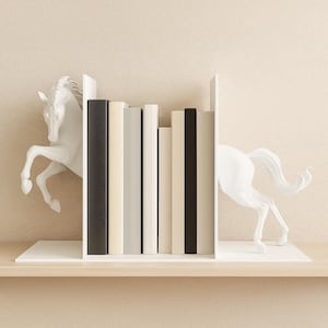 Puede incluir: Sujetalibros blancos con forma de caballo con una estantería que contiene una fila de libros. Los sujetalibros presentan un diseño de caballo encabritado. Los libros tienen varios colores, incluyendo negro, blanco y beige. La estantería es de madera.
