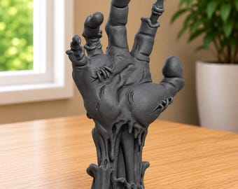 May include: A dark gray, three-dimensional sculpture of a severed hand with visible stitches, textured skin, and a melting or dripping base. The fingers are extended in an upright position, showcasing detailed anatomical features and a matte finish.