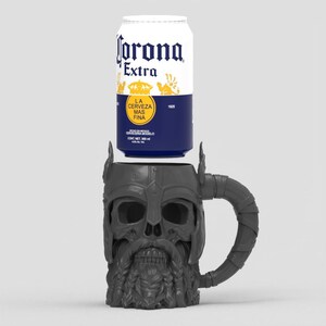 May include: A dark gray mug shaped like a bearded skull wearing a horned helmet, positioned directly beneath a 355 ml aluminum can of Corona Extra beer. The can features white, blue, and yellow branding with the text Corona Extra and La Cerveza Mas Fina.