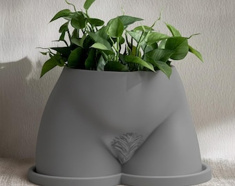 Könnte beinhalten: Grauer Keramik-Blumentopf in Form einer stilisierten weiblichen Figur, mit detaillierter Darstellung des Schambereichs. Der Topf ist mit grünen Blättern gefüllt. Der Topf steht auf einem passenden grauen Sockel.