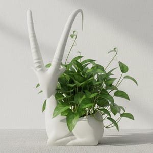 Puede incluir: Una maceta de cerámica blanca con forma de antílope, llena de plantas verdes vibrantes. El antílope tiene cuernos largos y curvos y está en posición sentada. Un elemento decorativo inspirado en la naturaleza.