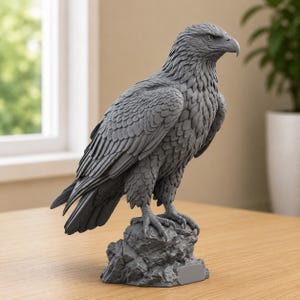 May include: A monochromatic grey eagle figurine standing on a textured rock base. The sculpture features detailed feather patterns across the wings and body, with the eagle looking towards the right. The base includes a small, blank rectangular plaque.