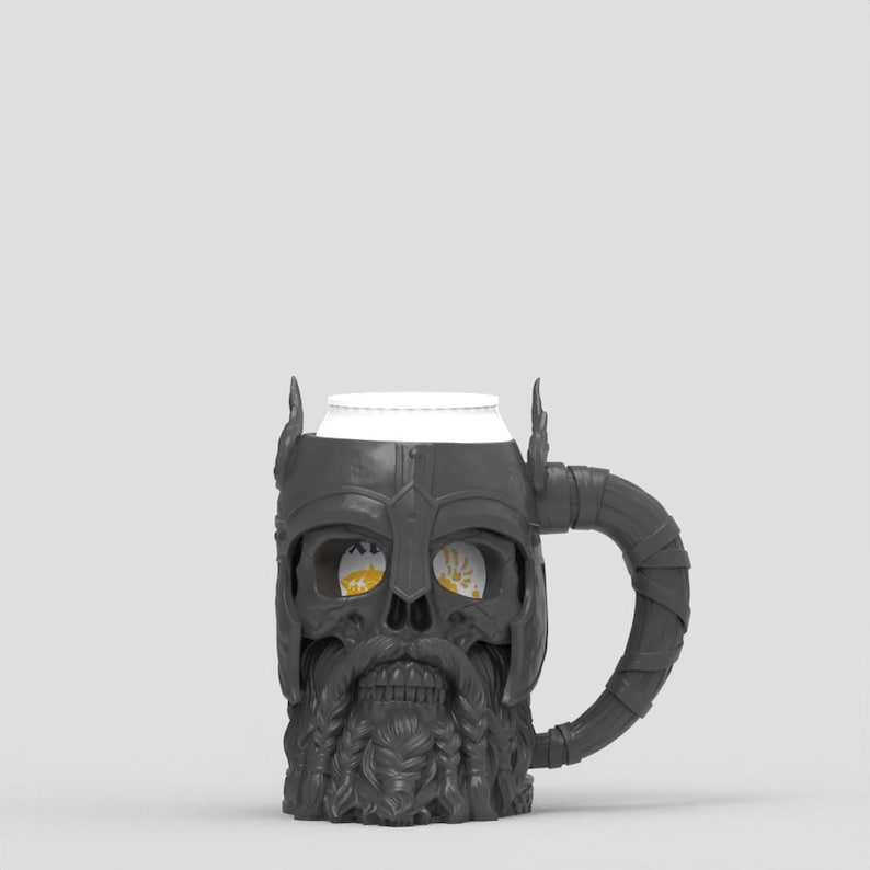 May include: A dark gray mug shaped like a bearded skull wearing a horned helmet. The mug features a textured, wrapped handle and a cylindrical white insert visible at the top. The eye sockets of the skull reveal a glimpse of a yellow graphic design.