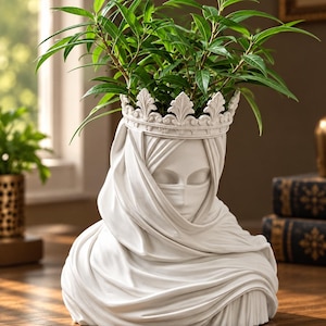 May include: A white ceramic planter shaped like a bust of a person wearing a head covering and a crown. The sculpture features detailed facial features, including eyes and a nose, with a draped fabric texture. A green leafy plant grows from the top.