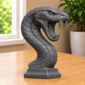 May include: A dark gray sculpture of a snake head with its mouth wide open, revealing sharp fangs and detailed scales. The serpent is depicted in a striking pose, rising from a square base, showcasing intricate textures and a matte finish throughout.