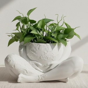 Puede incluir: Una maceta de cerámica blanca con forma de persona sosteniendo un globo, llena de plantas verdes. La maceta tiene tallas en relieve detalladas y está sobre una superficie neutra.