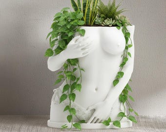 Könnte beinhalten: Ein weißer Keramik-Pflanztopf in Form eines weiblichen Torsos, verziert mit grünen Pflanzen und Ranken. Der Pflanztopf hat ein detailliertes Design mit Armen und einem spitzenbesetzten Kleidungsstück. Die Oberseite ist mit verschiedenen grünen Pflanzen gefüllt.