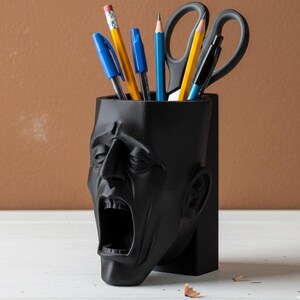 May include: A black desk organizer shaped like a screaming human head. The organizer holds pens, pencils, and scissors. The pencils are yellow with pink erasers, and the pens are blue and black. The scissors are black.