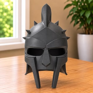 May include: A matte black gladiator-style helmet featuring a central vertical crest, a pointed brow ridge, and six small spikes protruding from the sides. The helmet has two large eye openings and a nose guard that extends down to the chin area.