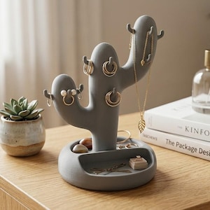 May include: Gray cactus-shaped jewelry holder with multiple hooks for hanging necklaces and earrings. The base has a small tray for rings and other small accessories. A succulent plant in a small pot is to the left of the jewelry holder.