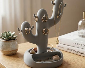May include: Gray cactus-shaped jewelry holder with multiple hooks for hanging necklaces and earrings. The base has a small tray for rings and other small accessories. A succulent plant in a small pot is to the left of the jewelry holder.