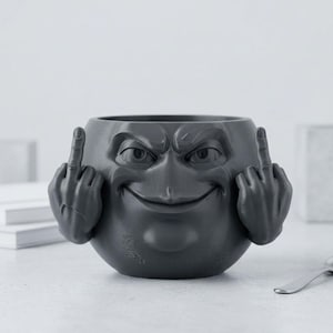 Puede incluir: Un cuenco de cerámica gris oscuro con un diseño de rostro, con una amplia sonrisa, cejas fruncidas y ojos. El cuenco tiene dos manos a cada lado, cada una mostrando el dedo medio. El cuenco está sobre una superficie blanca.