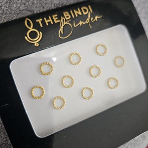 Könnte beinhalten: Eine schwarze Box mit dem Text "The Bindi Binder" auf der Oberseite. Die Box enthält 10 goldfarbene runde Bindis.