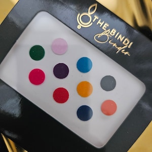 Può includere: Un set di dieci bindis rotondi colorati in una custodia di plastica bianca con il testo "The Bindi Binder" stampato sul coperchio nero.