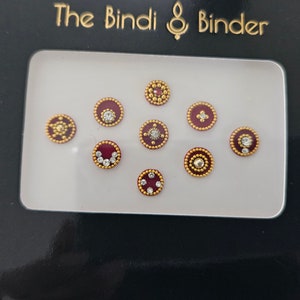 May include: A set of ten red and gold bindis with various rhinestone designs. The bindis are packaged in a clear plastic case with a black backing card that reads "The Bindi & Binder".