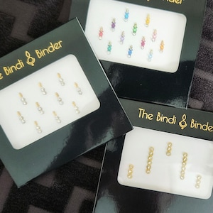Può includere: Tre scatole di imballaggio in cartone nero con il testo "The Bindi & Binder" sul davanti. Ogni scatola contiene un set diverso di piccoli bindis adesivi trasparenti con accenti dorati.