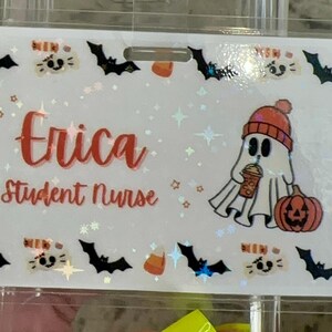 Pode incluir: Um crachá de plástico branco com fundo brilhante. O crachá tem um fantasma segurando uma xícara de café e uma abóbora. O texto "Erica Student Nurse" está impresso em vermelho no crachá.