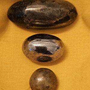 May include: Three smooth, round, metallic stones of varying sizes. The largest stone is oval-shaped and the smallest stone is spherical. The stones are arranged vertically on a yellow fabric background.