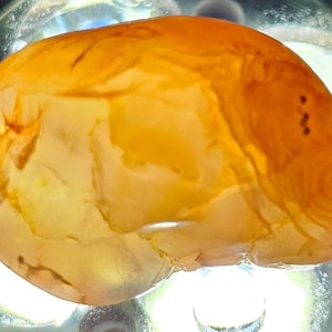 May include: A smooth, orange and white agate stone with a natural, organic shape. The stone has a translucent quality and shows intricate patterns of colour.