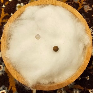 Puede incluir: Dos pequeñas cuentas redondas, una blanca y otra marrón, están anidadas en un lecho de fibras de algodón blancas dentro de un cuenco de madera.