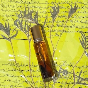 Könnte beinhalten: Eine kleine, klare Glas-Parfümflasche mit goldenem Deckel, gefüllt mit einer dunkelbernsteinfarbenen Flüssigkeit. Die Flasche steht vor einem gelben Hintergrund mit handschriftlichem Text und Pflanzenillustrationen.