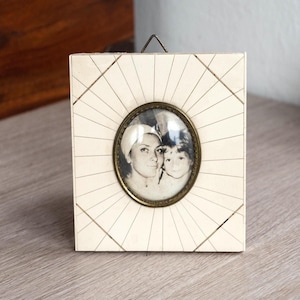 May include: A square, off-white wooden picture frame featuring a sunburst pattern of thin, etched lines radiating from a central oval opening. The frame contains a black and white photograph of two people, enclosed by a dark metallic oval border.