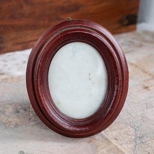 Pode incluir: Uma pequena moldura oval de madeira vermelha escura. A moldura tem um design em camadas e uma pequena argola de metal na parte superior para pendurar. O interior contém um espaço oval esbranquiçado desbotado para uma foto ou obra de arte.