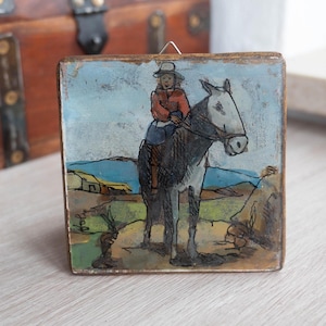 Könnte beinhalten: Ein kleines, quadratisches, vintage Gemälde zeigt eine Person auf einem Pferd in einer Landschaft. Der Reiter trägt eine rote Jacke und einen Hut. Das Pferd ist grau, und der Hintergrund zeigt einen blauen Himmel und grüne Hügel. Das Gemälde ist auf einem Holzblock montiert.