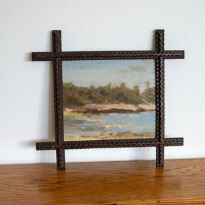 May include: A small landscape painting in a dark brown, intricately carved wooden frame. The painting depicts a coastal scene with a blue sky, water, and green trees. The frame has a cross-like design at each corner.