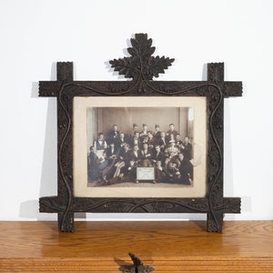 Pode incluir: Uma moldura de madeira marrom escura, ornamentada e esculpida, com uma fotografia em preto e branco de um grupo de pessoas. A moldura tem detalhes florais e de folhas esculpidas, e um grande desenho de folha no topo. A foto mostra homens em trajes formais.