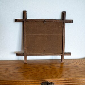 May include: Back of a wooden picture frame with a dark brown backing. The frame is made of dark wood with a cross-like design on each corner. A small metal hook is attached to the top center for hanging. The frame is sitting on a wooden surface.