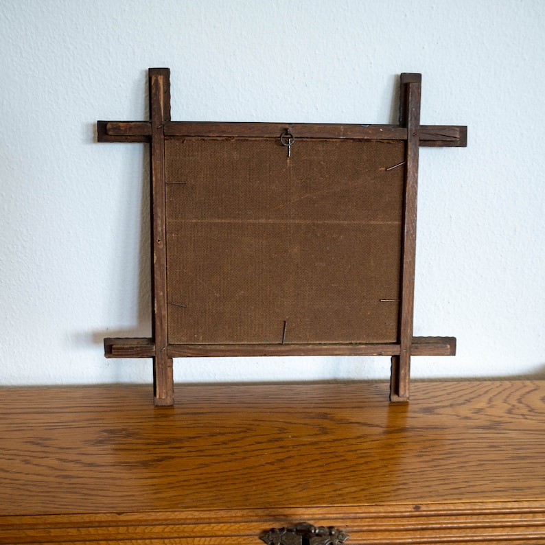 May include: Back of a wooden picture frame with a dark brown backing. The frame is made of dark wood with a cross-like design on each corner. A small metal hook is attached to the top center for hanging. The frame is sitting on a wooden surface.