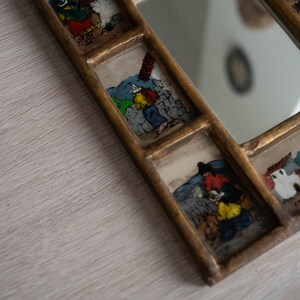 May include: A gold framed mirror with multiple small glass panels featuring hand-painted scenes of people in various settings. The mirror is on a light wood surface.