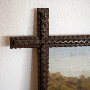 May include: Close-up of a rustic, cross-shaped picture frame. The frame is made of dark, textured wood with a decorative, notched edge. The painting within the frame depicts a landscape with trees and a cloudy sky.