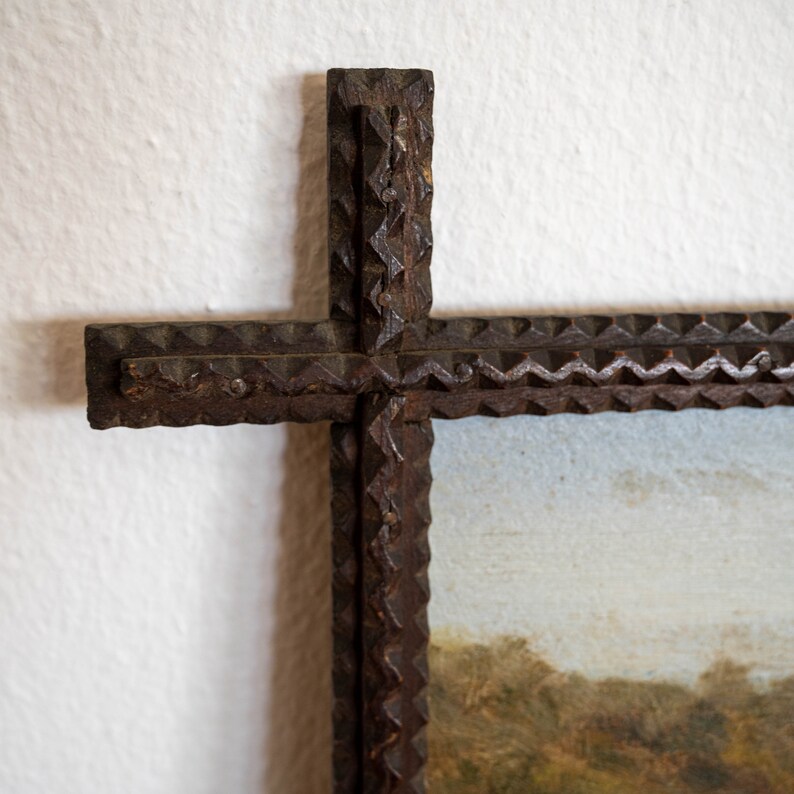 May include: Close-up of a rustic, cross-shaped picture frame. The frame is made of dark, textured wood with a decorative, notched edge. The painting within the frame depicts a landscape with trees and a cloudy sky.